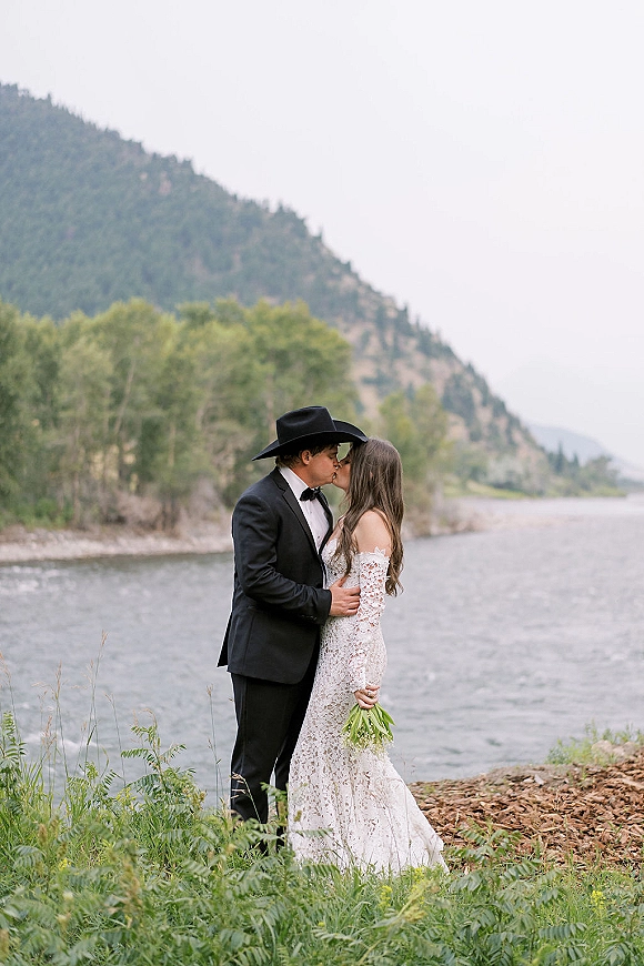 Wedding kiss portrait of bride and groom kissing, her lace off-shoulder dress and green bouquet beside a mountain lake shoreline with forest trees