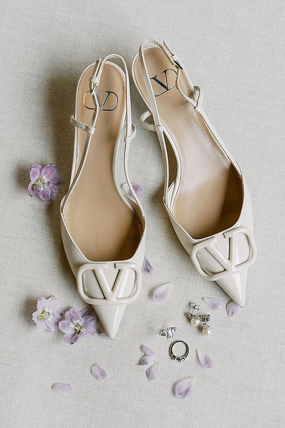 Bridal shoes in a flat lay with white slingback heels on linen, paired with wedding rings, pearl drop earrings, and purple petals