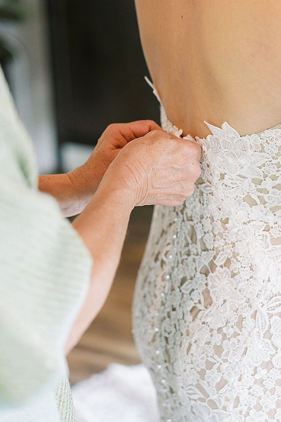 Wedding dress buttoning as hands fasten tiny back buttons on a lace gown with beaded applique, in an indoor room with a blurred helper behind