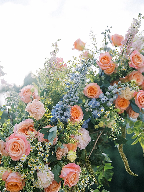 Wedding floral arrangement with peach rose wedding flowers, blue delphinium and eucalyptus, set against sunlit sky and outdoor foliage