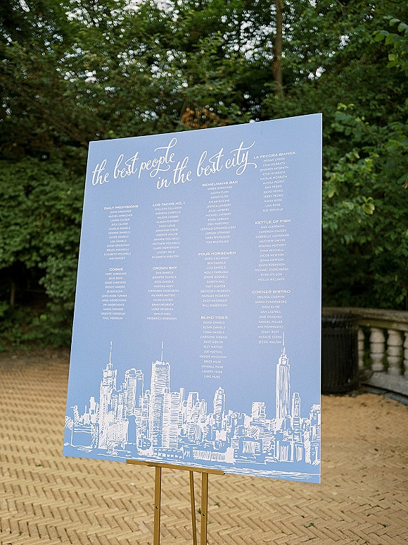 Wedding seating chart sign with calligraphy lettering and illustrated skyline on an easel, set on a brick patio with trees behind