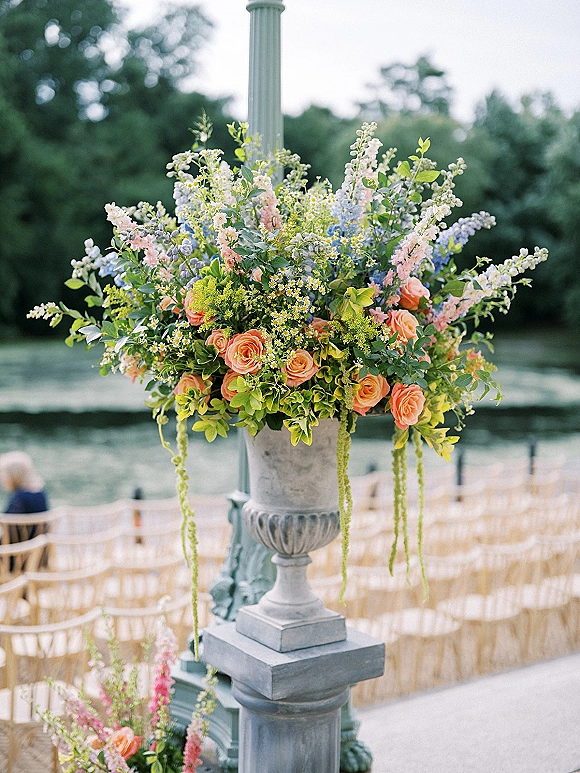 Ceremony floral arrangement in a stone urn with pastel wildflowers, peach roses, and trailing amaranthus by a lakefront aisle of chairs