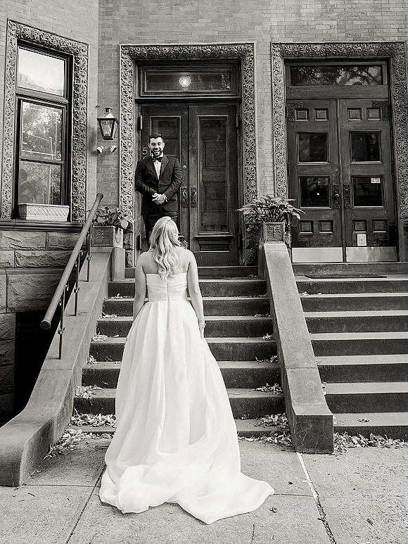 First look moment as bride approaches groom on stone steps, her strapless gown’s long train flowing beside a brick doorway