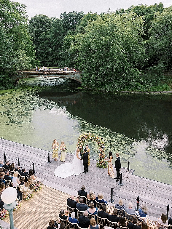 Outdoor wedding ceremony at a waterfront deck with floral arch, bride and groom at altar, guests seated by pond and bridge.
