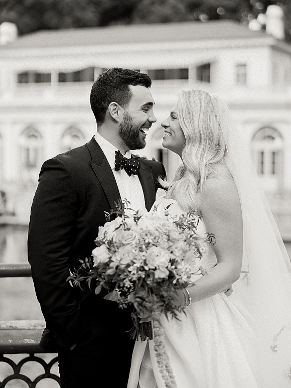 Couple portrait in a black and white wedding portrait style, bride and groom smiling face to face on a balcony, veil and bouquet visible
