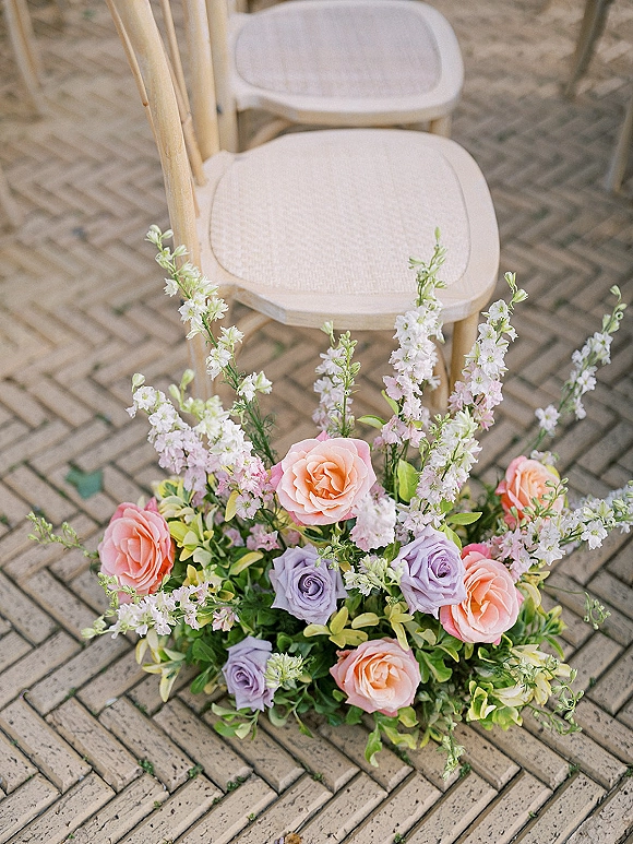 Aisle floral arrangement of pastel and lavender roses with tall white blooms and greenery beside a wooden chair on a brick patio