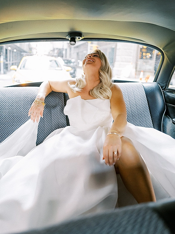 Bridal portrait of a bride in car, laughing in a strapless wedding dress with gold bracelet and ring, wavy hair, city street beyond window