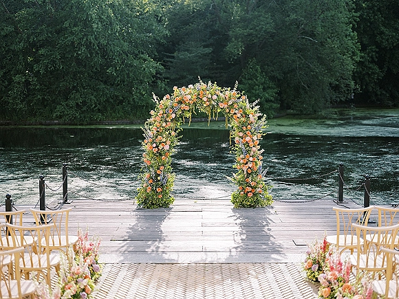 Wedding ceremony arch with roses and greenery on a sunlit dock, aisle flowers and wooden chairs facing a lake and trees