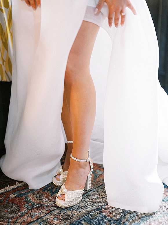 Bridal shoes, pearl wedding heels with ankle strap and open toe, shown as bride adjusts them beside her ring on a vintage rug indoors