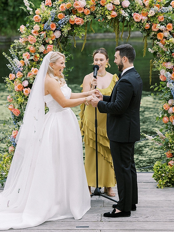 Ceremony moment as bride and groom hold hands under a rose floral arch on a lakeside dock, officiant speaking by a microphone stand
