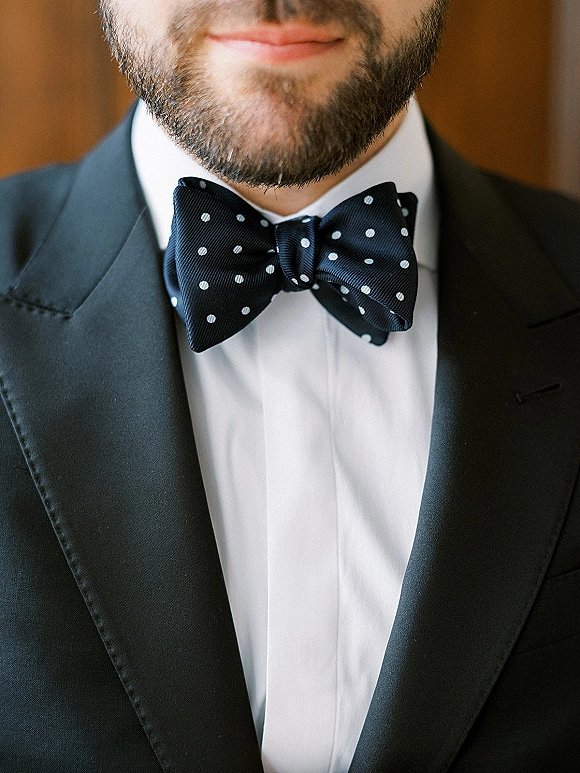 Groom bow tie with polka dot bow tie centered on a bearded groom in a black tuxedo jacket with satin lapel and white shirt indoors