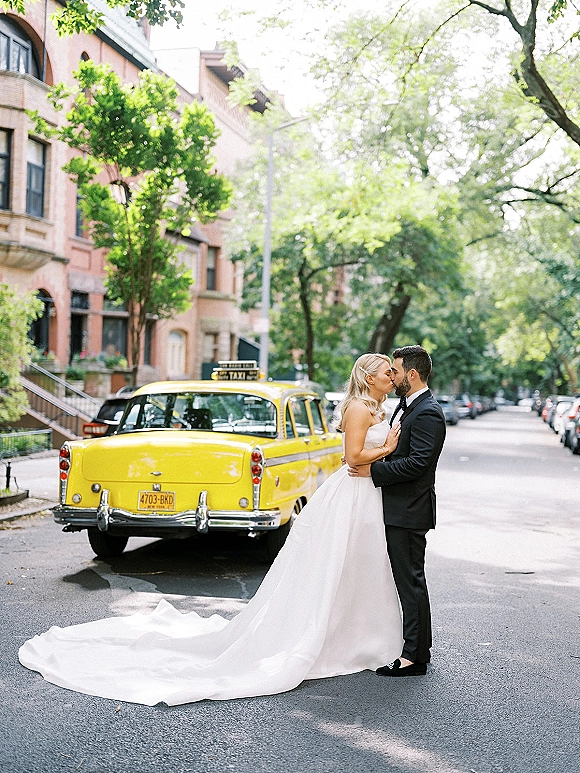 Wedding kiss portrait of bride and groom kissing in front of a vintage yellow taxi, her strapless gown’s long train on a tree-lined city street