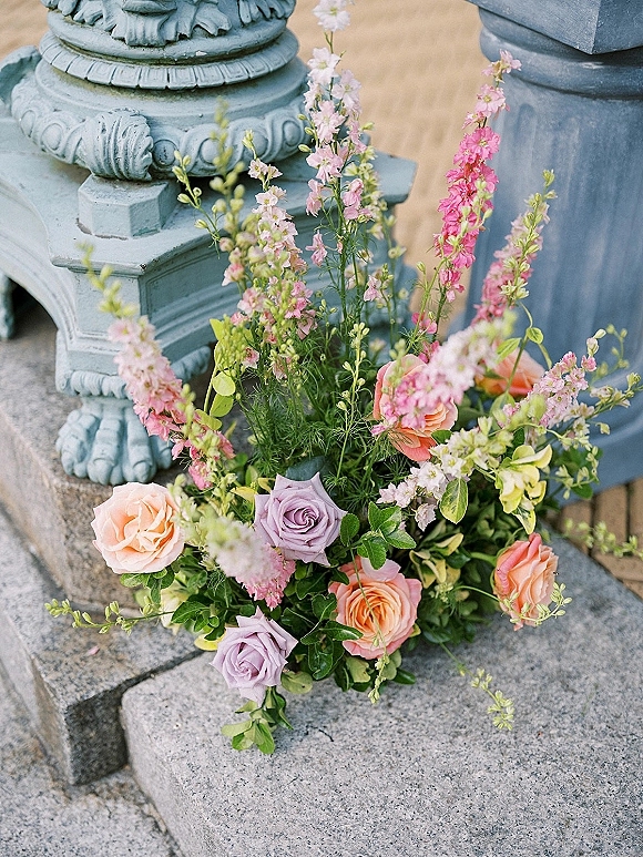 Wedding floral arrangement with peach and lavender roses spilling from a stone urn on stone steps with greenery accents on patio