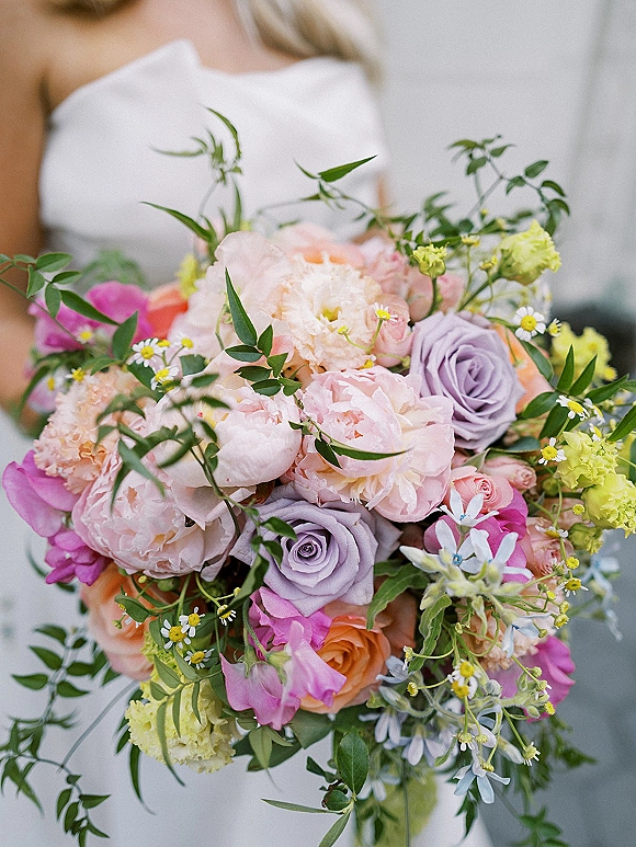 Bridal bouquet in a pastel wedding bouquet style with peonies, lavender and coral roses, chamomile daisies, and ribbon against a white dress