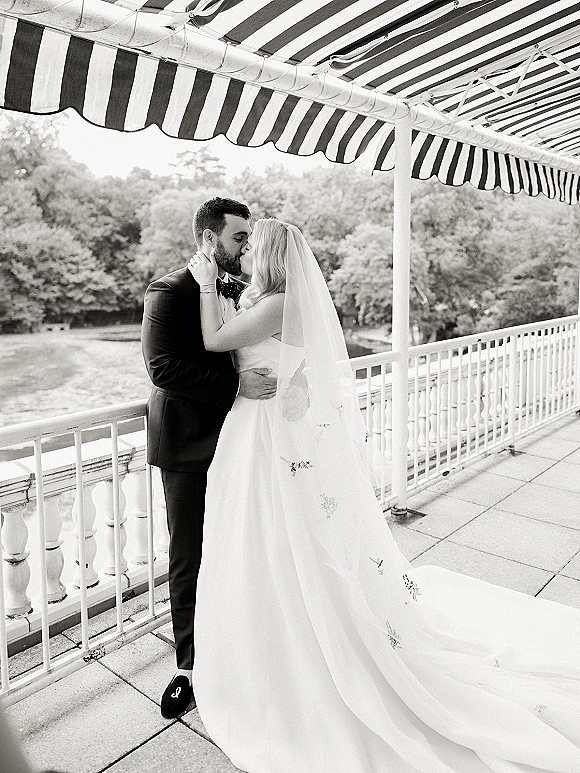 Wedding kiss portrait of bride and groom kissing under a striped awning on a terrace, her embroidered veil and long train flowing