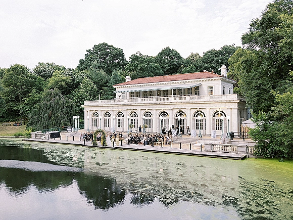 Outdoor ceremony setup with lakefront wedding ceremony chairs lining an aisle to a floral arch on a stone terrace by the dock and water