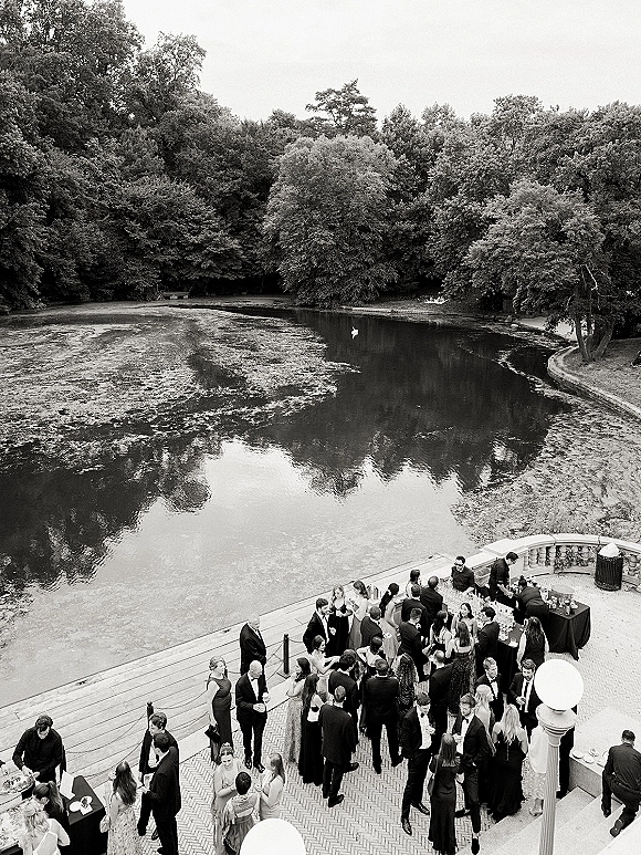 Wedding cocktail hour with guests mingling in outdoor cocktail hour attire around cocktail tables and bar setup on a lakeside terrace with globe lamps