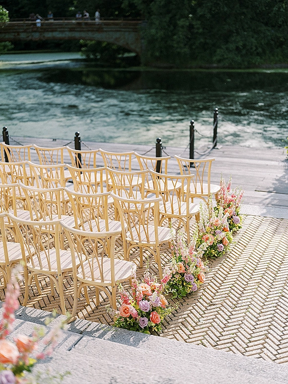 Ceremony seating setup with outdoor ceremony chairs in a curved layout, lined by rose and greenery florals on a riverside terrace with stone bridge