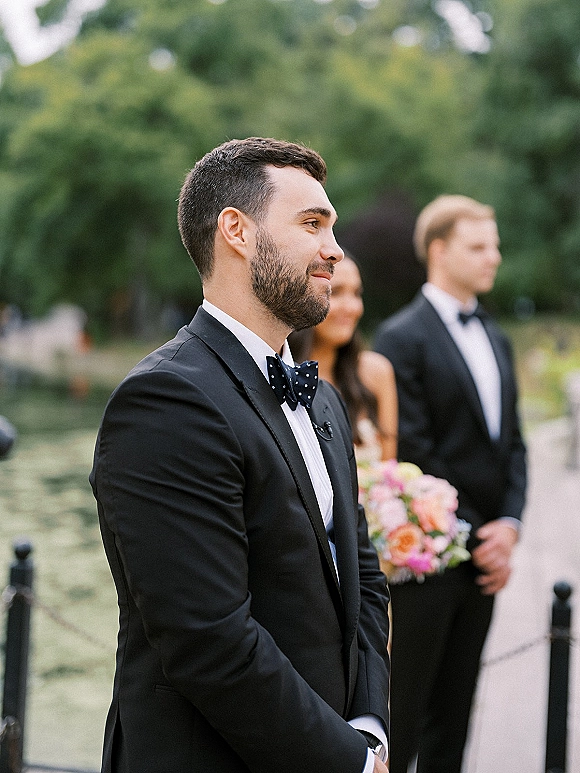 Groom portrait in a black tuxedo with polka dot bow tie and boutonniere, checking his wristwatch by a garden pond walkway.