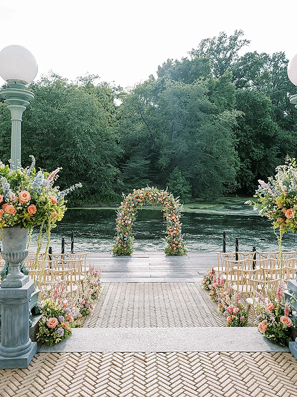 Ceremony setup for an outdoor wedding ceremony with a floral arch and rose-lined aisle on a lakefront terrace with stone steps and wooden chairs