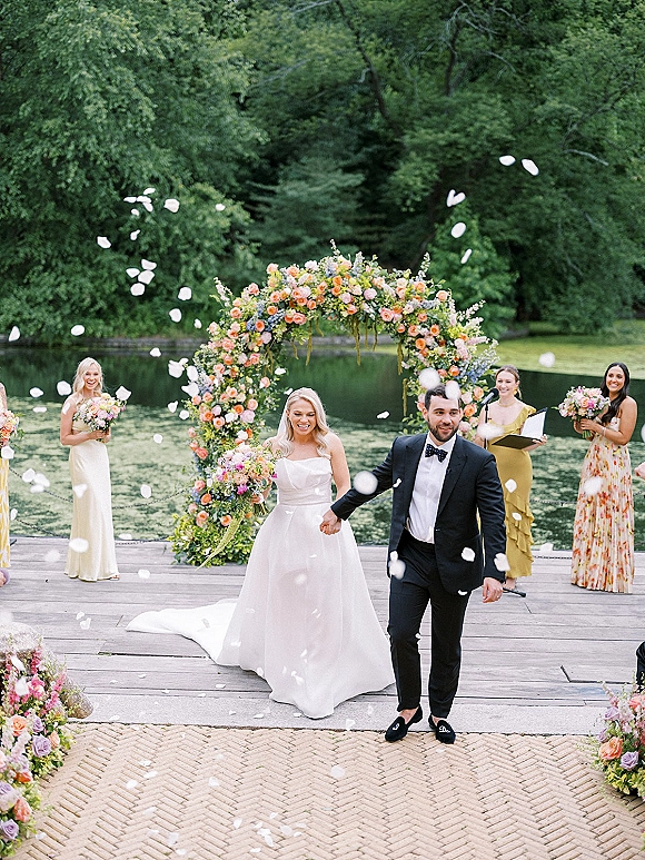 Wedding recessional as newlyweds walk hand in hand down the aisle, bride holding bouquet, guests tossing flower petals on a lakeside dock