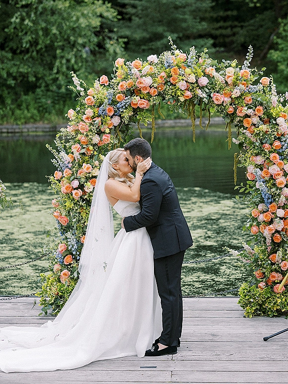 Wedding kiss as bride and groom embrace under a rose-and-greenery floral arch on a wooden dock by a lake, veil trailing behind
