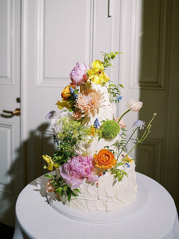 Wedding cake with textured buttercream frosting and fresh flowers on a cake stand, set by white paneled doors with sunlit shadows