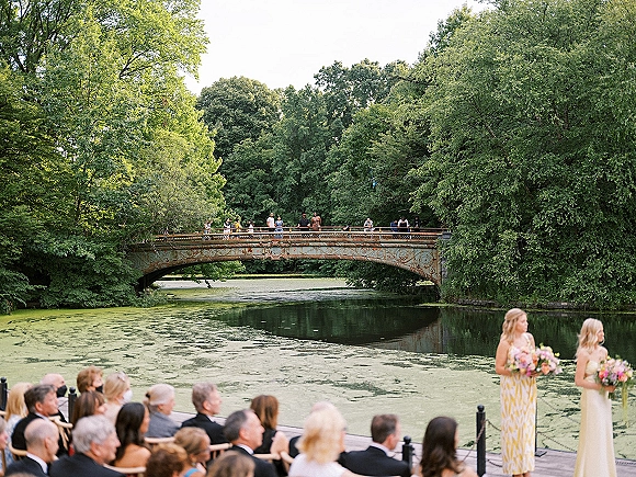 Outdoor wedding ceremony by a pond with guests seated, bridesmaids holding colorful bouquets, and a bridge framed by trees in back