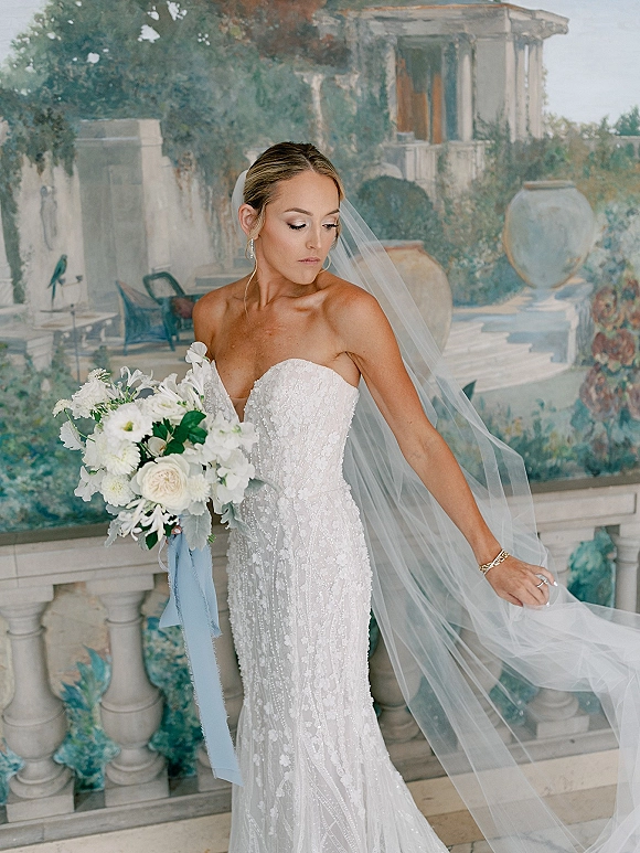 Bridal portrait of a bride in a strapless lace wedding dress with long veil, holding a white bouquet with blue ribbon by a mural terrace balustrade