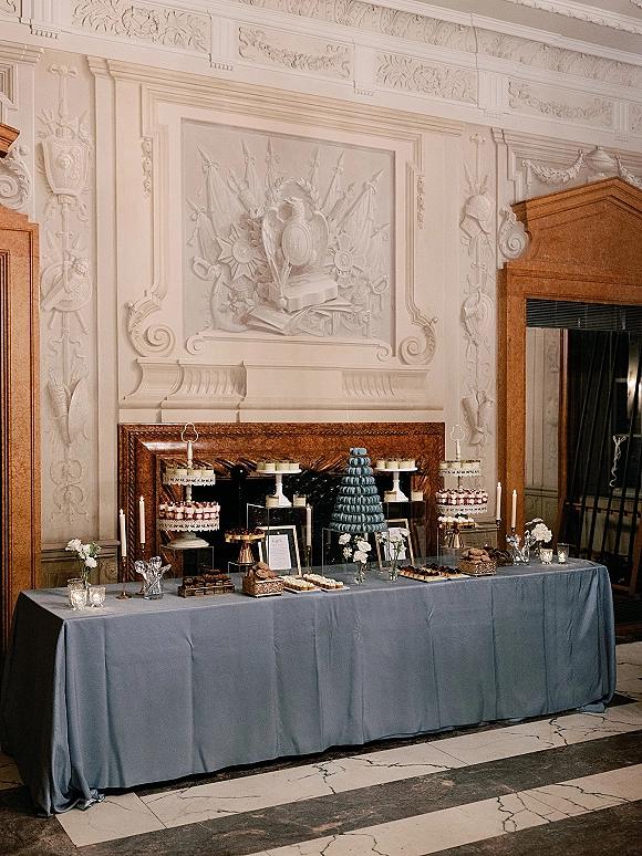 Wedding dessert table with macaron tower wedding display, tiered stands, mini cakes, candles and signage before an ornate fireplace wall relief