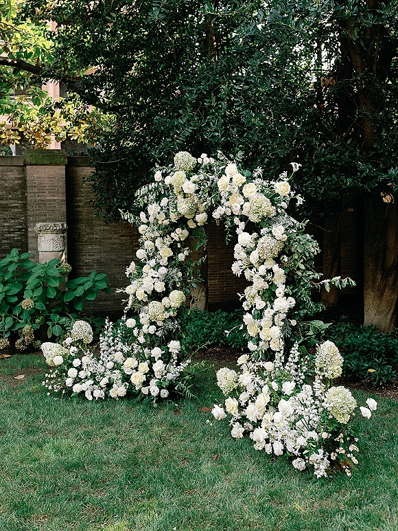 Wedding flower arch with white roses and hydrangeas, lush greenery, and ground blooms on a garden lawn beside trees and a brick wall