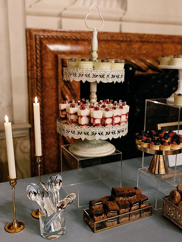 Wedding dessert table with a tiered dessert stand, mini dessert cups, berry tarts, and taper candles before an ornate framed mirror
