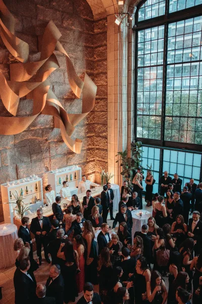 Wedding cocktail hour with guests mingling in an indoor hall; champagne bottles gleam as tall windows reveal city buildings against a stone wall.