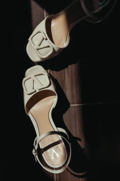 Bridal shoes, ivory bridal heels with subtle logo hardware, rest on a sunlit wood floor, their clean lines suited for a modern bride.