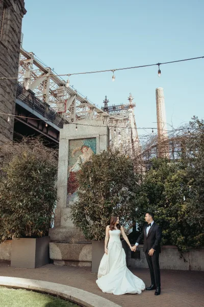 Couple portrait of a bride and groom holding hands, facing each other under string lights with a bridge structure and mosaic panel backdrop.