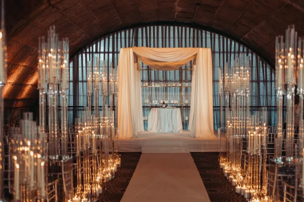 Ceremony setup featuring a candlelit ceremony aisle under arched windows, with a vaulted ceiling and stone walls framing a gold frame arch.