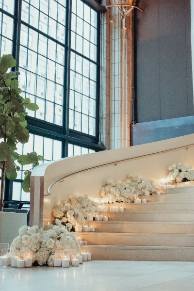 Staircase wedding decor lines a candlelit staircase, with white florals clustered on each step beside industrial windows and a tile floor.