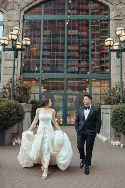 Couple portrait of bride and groom walking, her strapless bridal gown swaying beside him under string lights in urban courtyard.