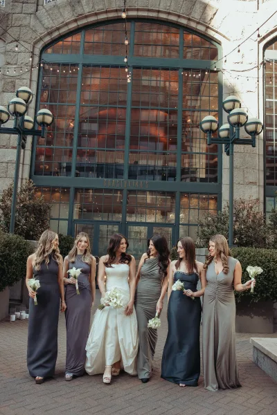 Bridal party portrait of a bride with bridesmaids carrying calla lily bouquets in a paved courtyard beside industrial windows under string lights.