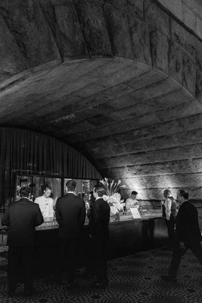 Wedding bar setup for a cocktail hour bar beneath a stone arch with drapery, featuring a refined floral arrangement along the bar.