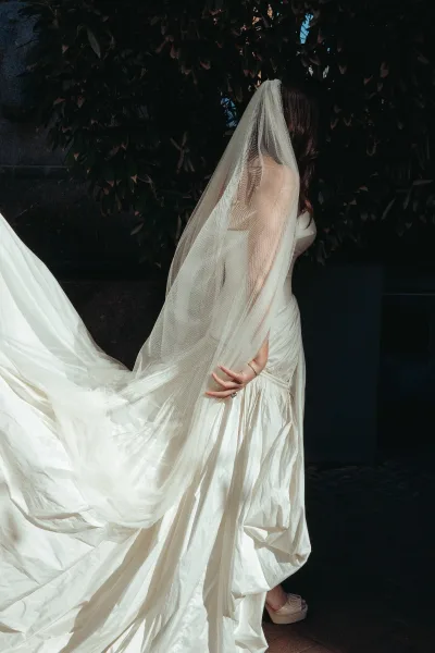 Wedding dress detail of a bride with veil, showcasing an ivory strapless gown and flowing train beside a sunlit stone wall.