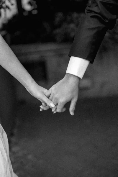 Couple holding hands with interlocked fingers, a close-up showing the groom’s white shirt cuff as they stand over dark stone paving.