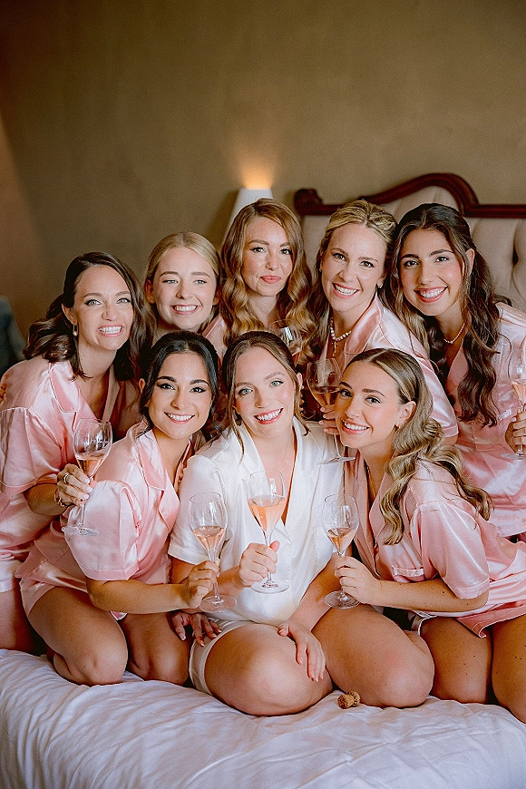 Bridesmaids getting ready in matching pink satin pajamas with champagne flutes, gathered on a hotel bed beneath a neutral wall
