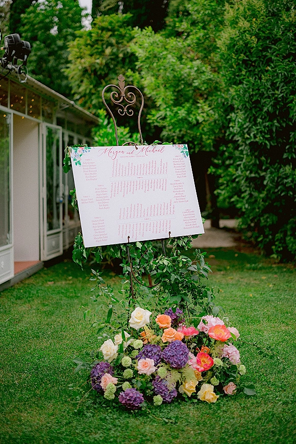 Wedding seating chart on a wrought iron easel with greenery garland and roses, set on a lawn with greenhouse behind