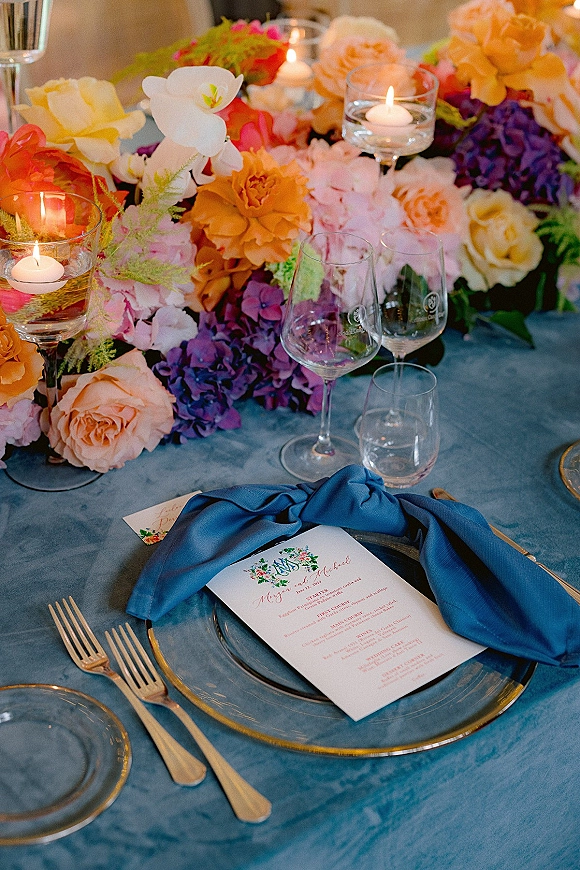 Reception tablescape with a wedding place setting, colorful floral centerpiece, floating candles, blue velvet napkin, and gold flatware on blue tablecloth