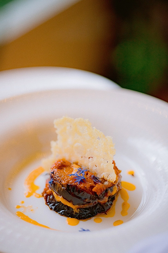 Wedding plated dinner appetizer in a white bowl with sauce drizzle, edible flower accents, and crispy garnish on a blurred table setting