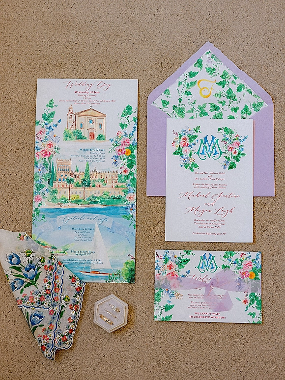 Invitation suite with watercolor wedding invitation, floral stationery and monogram crest beside rings and lavender envelope on beige carpet