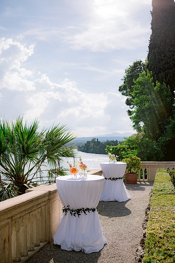 Cocktail hour setup with wedding cocktail tables in white linens, small floral arrangements in glass vases on a stone terrace overlooking a lake and mountains