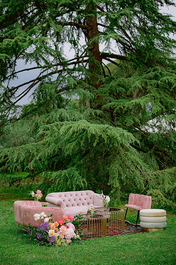 Wedding lounge seating with a pink velvet sofa and chairs around gold coffee tables, styled with florals on a patterned rug on a green lawn