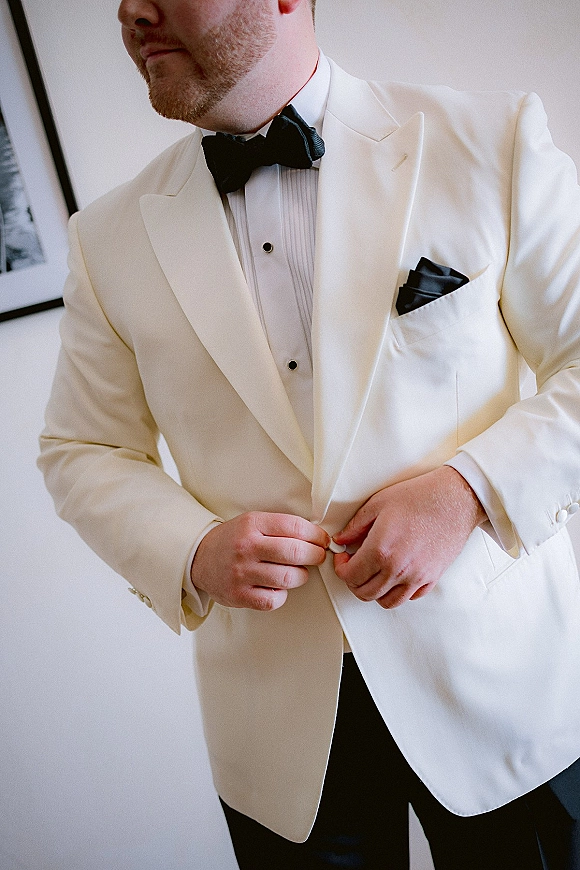 Groom getting ready, buttoning a white tuxedo jacket with black bow tie and pocket square against a white wall with framed art